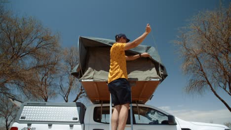 wide static shot of a off-roading caucasian male tourist in africa standing on the ladder of a rooftop tent while he inserts a peg to the tent door.