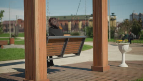 mujer en una silla de columpio balanceándose con el cabello revoloteando en el viento, el fondo de árboles y personas borrosas sentadas en la distancia en un parque público, capturando la relajación al aire libre serena