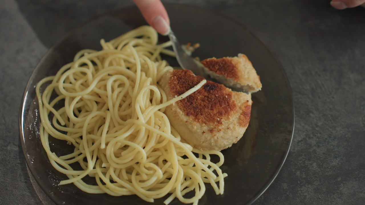 Piatto di spaghetti e cotoletta impanata servito, primo piano dall'alto di un semplice pasto fatto in casa su un piatto di vetro, forchetta e cucchiaio che porzionano la cotoletta, luce serale soffusa sul bancone della cucina, atmosfera accogliente e minimale.