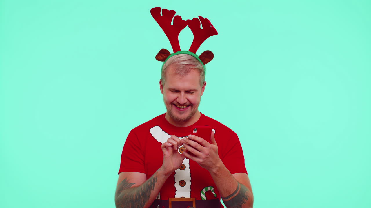 hombre con camiseta de navidad usando teléfono móvil escribiendo una nueva publicación en la web, haciendo compras en línea, mostrando el pulgar hacia arriba