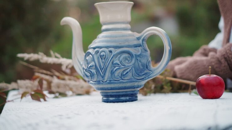 Blue ceramic teapot on tablecloth, red apple beside ornate porcelain vessel, dried foliage and knit sleeve of person arranging scene, outdoor autumn garden light, shallow depth of field, warm cozy