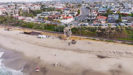 Aerial-View-of-California-beach-town-with-surfers-crossing-bridge-over-train-tracks