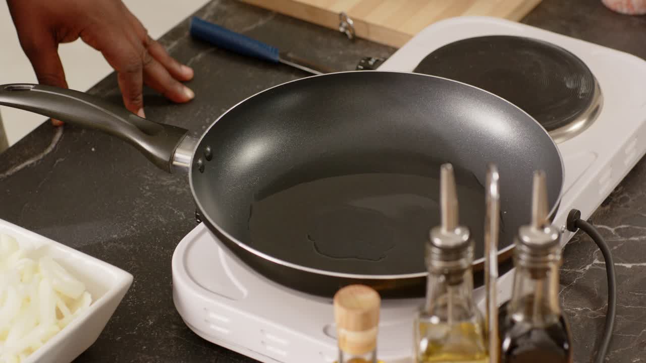 Un uomo africano sta inclinando una padella su una piastra calda bianca riscaldando l'olio, lasciando cadere anelli di cipolla nell'olio sul bancone.