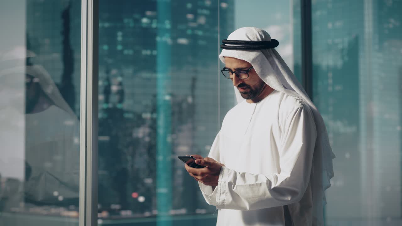 Successful Muslim Businessman in Traditional White Outfit Standing in His Modern Office, Using Smartphone Next to Window with Skyscrapers. Successful Saudi, Emirati, Arab Businessman Concept.