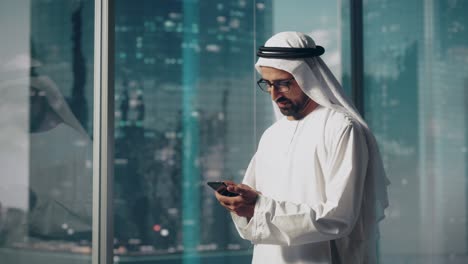 successful muslim businessman in traditional white outfit standing in his modern office, using smartphone next to window with skyscrapers. successful saudi, emirati, arab businessman concept.