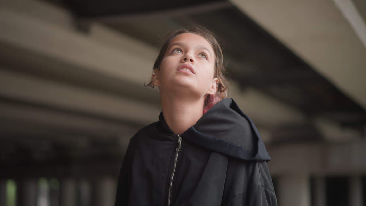 Young woman under overpass in rain, black hooded coat, raindrops in hair, looking up at sky, stepping around puddles, quiet contemplative mood with concrete columns and muted city light