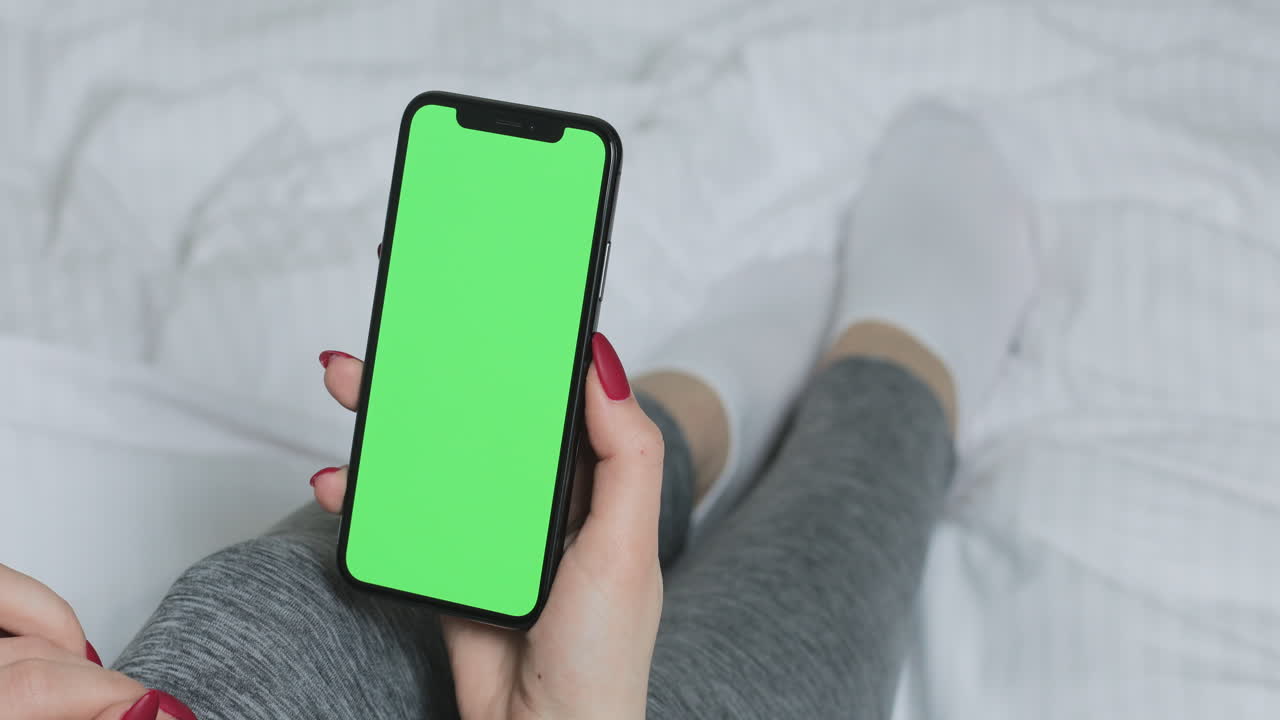 Woman holding a smartphone with a green screen on a bed