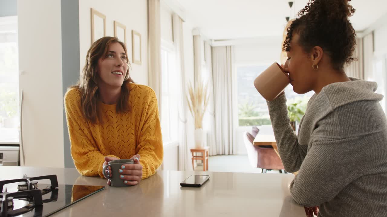 Diverse female friends resting mugs beside smartphone, engaging in friendly chat at kitchen island