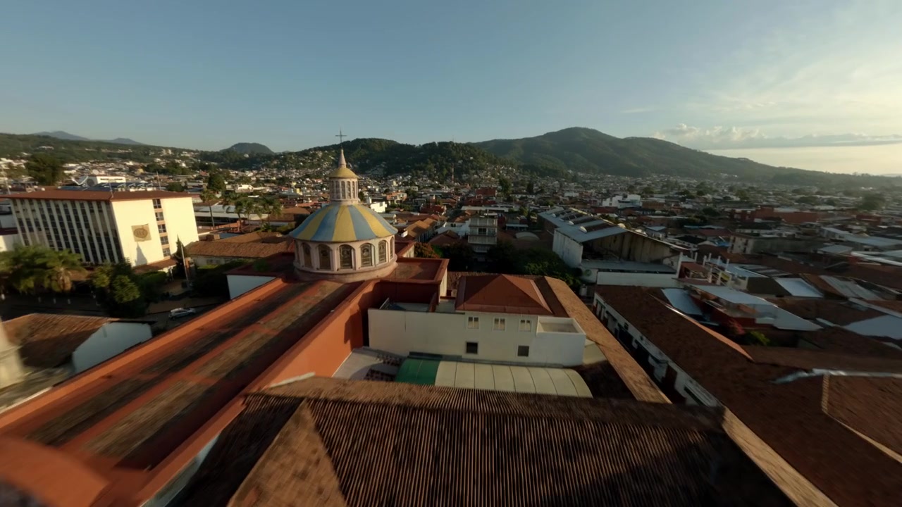 Sorvolo aereo dinamico FPV di una cupola di chiesa e di un paesaggio urbano con palme durante l'ora d'oro, Uruapan, Michoacán, Messico.