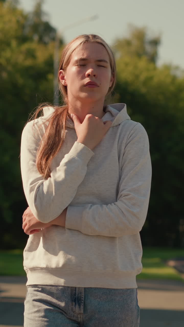 Young woman with arms folded, thoughtfully touching her hoodie, walking outdoors near stadium, background includes trees, electric poles, and stadium seating