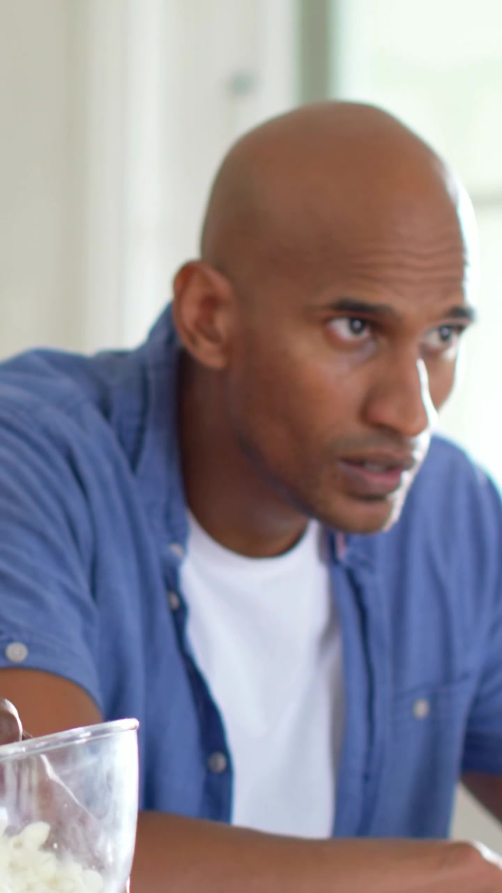Vertical video: Leaning man in blue shirt moving green bottle and replying offscreen voice at table