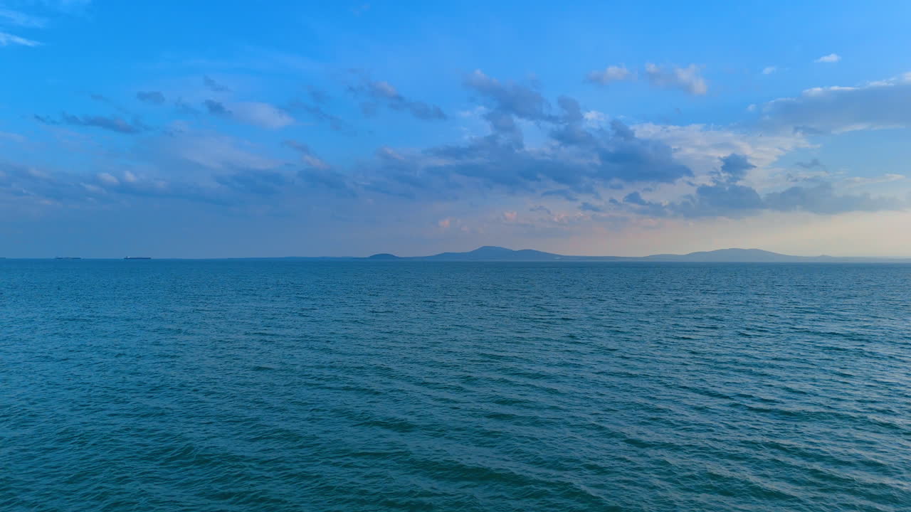 Acque calme e azzurre del mare sotto un cielo nuvoloso, vicino a colline. Vista grandangolare della superficie dell'oceano con montagne lontane sotto un cielo azzurro.