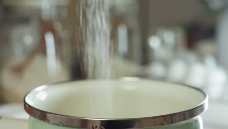 close up of flour being poured in steady stream into metal-rimmed white pot during food preparation, with soft background blur showing eggs and kitchen setup in natural light for cooking process