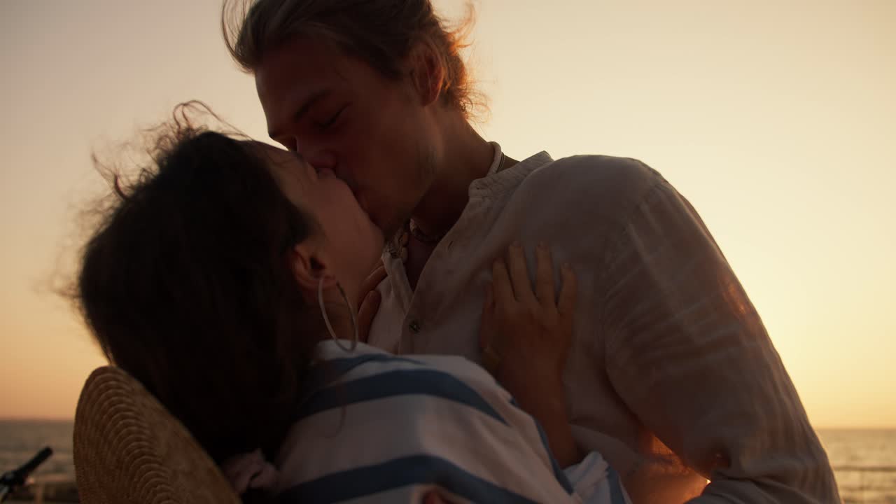 Happy couple, a guy in light clothes and a girl in a white and blue shirt kissing at Sunrise near the sea. Happy couple on a date on the beach near the sea at sunrise in summer