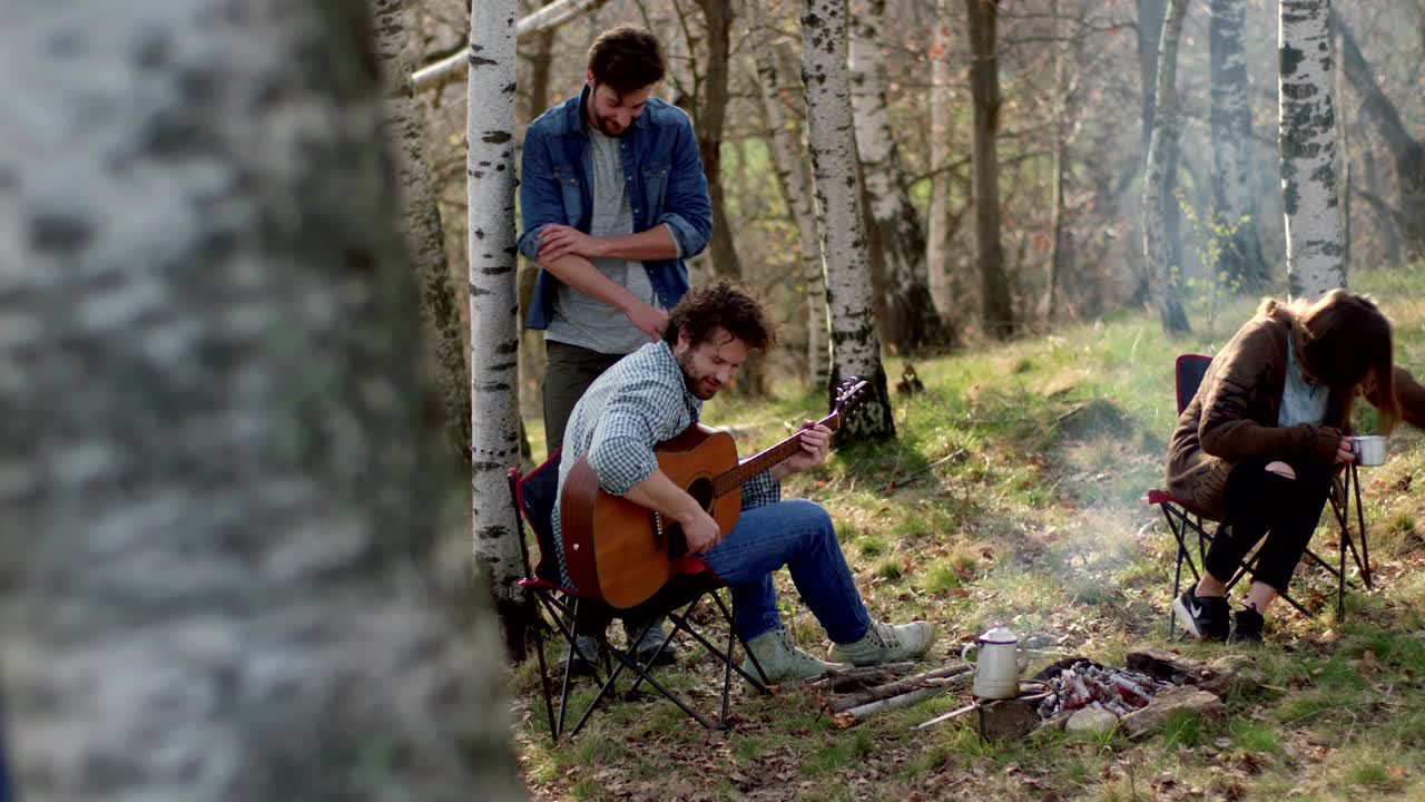 hombre tocando la guitarra y la mujer acariciando al perro juntos alrededor de la chimenea. verdaderos amigos personas al aire libre acampando vacaciones en la tienda de campaña en el viaje de otoño. caída día soleado en la naturaleza, unión y amistad. video 4k