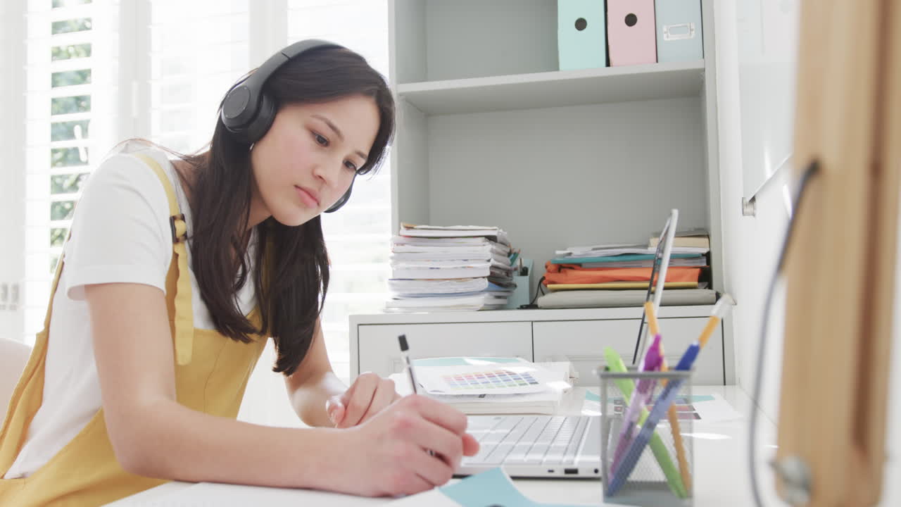 두 인종의 여성이 헤드폰을 착용하고 노트북을 사용하고 책상에 앉아서 집에서 일하고, 느린 동작