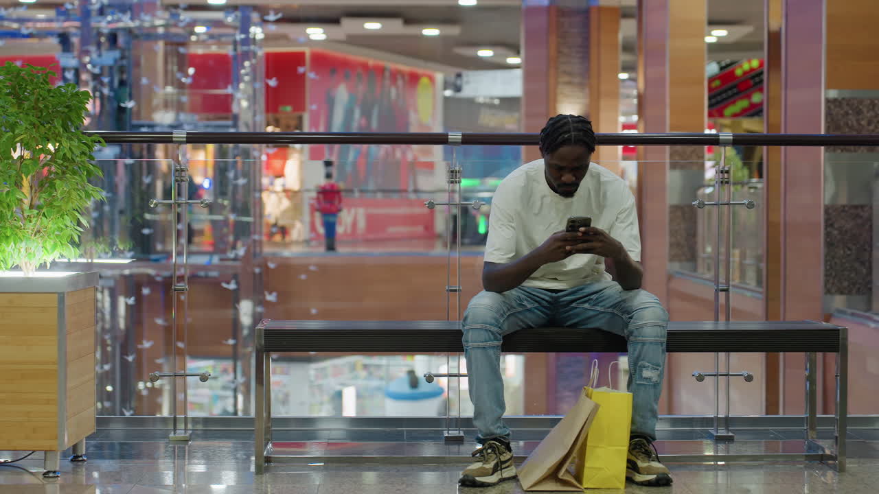 Black man sitting on bench using smartphone while looking around in brightly lit shopping mall with vibrant interior, surrounded by glass walls, green plant, and paper shopping bags on shiny floor