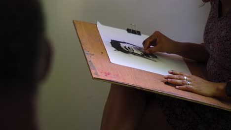 drawing class close up on hands of an olive skin woman wearing silver rings and drawing amateur portrait learning how to in art class not sure what she is doing sitting inside dark room natural light