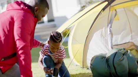 Happy-african-american-father-and-son-pitching-tent-together-in-sunny-garden,-in-slow-motion