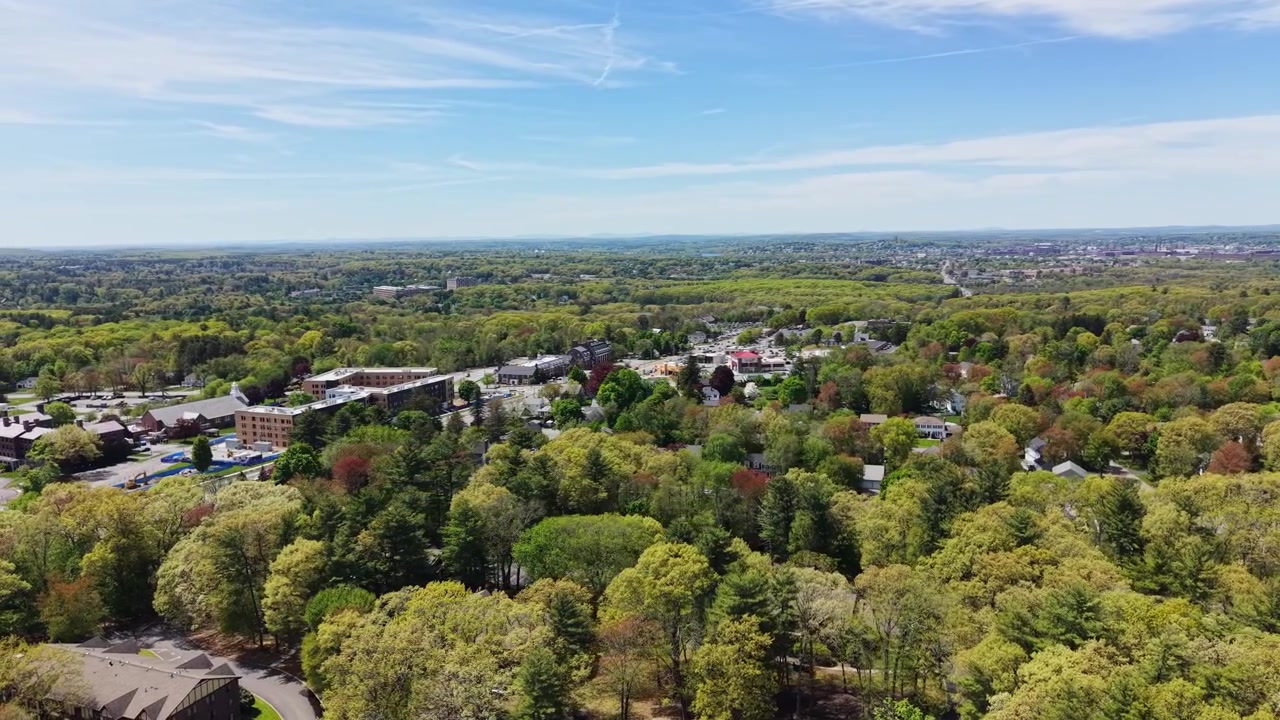 Un exuberante dosel de árboles de verano cubre un tranquilo paisaje suburbano con edificios de ladrillo y tejados asomándose a través de un denso bosque verde en North Andover, Massachusetts, EE. UU. Plano general aéreo con dron