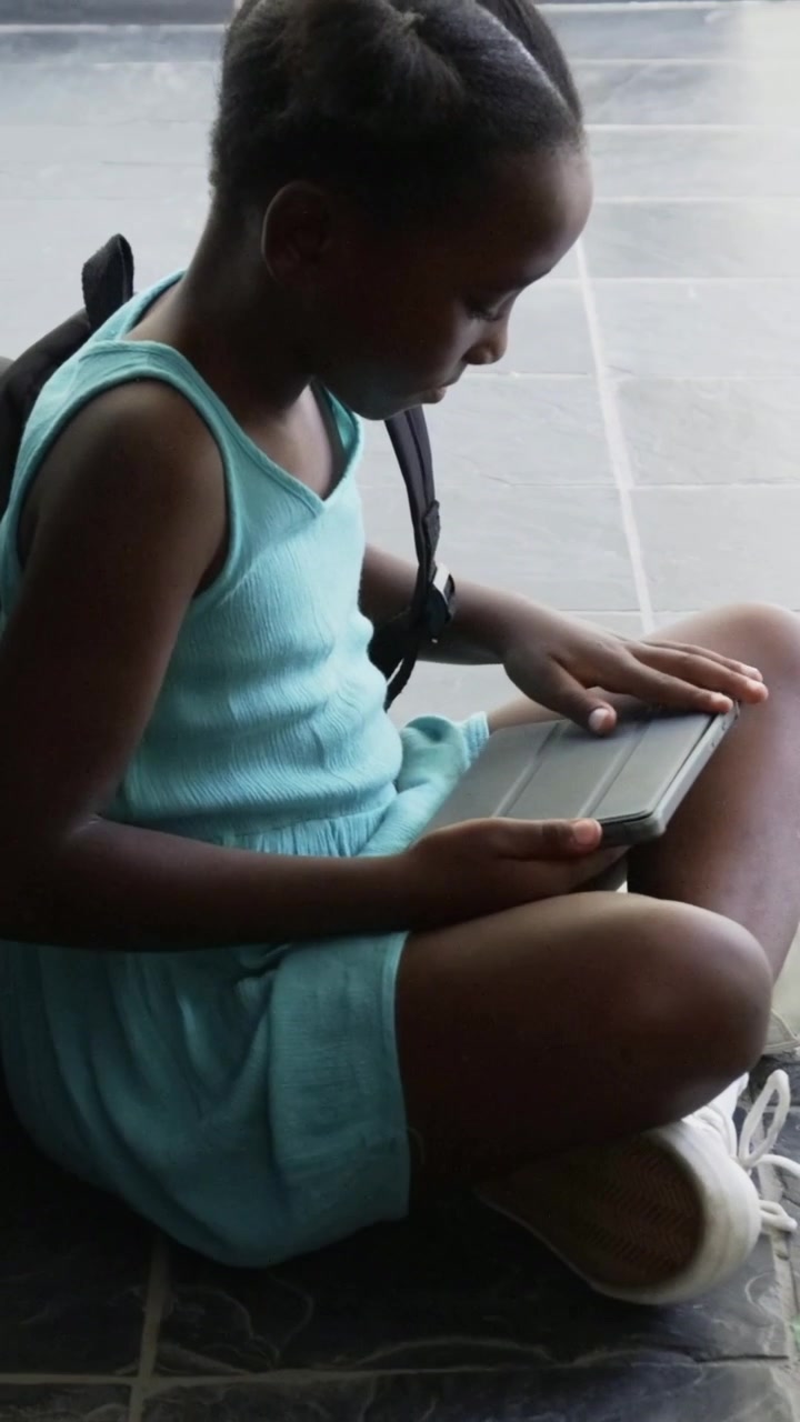 Vertical video: Shifting weight girl sitting on tile floor, tapping tablet waiting in blue dress