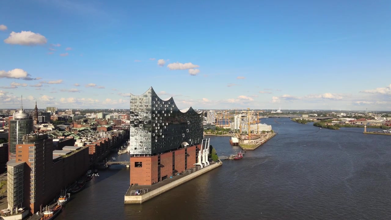 Ripresa aerea con drone di una barca che naviga lungo il fiume Elba, oltre l'Elbphilharmonie, con edifici storici e moderni sul lungofiume e gru portuali ad Amburgo, Germania.