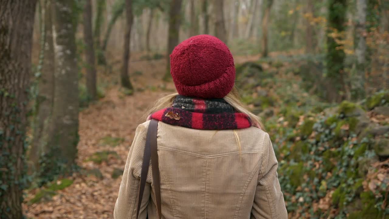 Ripresa da dietro di una donna con cappello rosso che cammina su un sentiero in un bosco autunnale, Slow motion