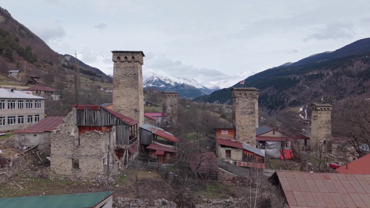 Vista aérea de drone exibindo torres Svan medievais com a bandeira georgiana tremulando, emolduradas pelas deslumbrantes montanhas nevadas do Cáucaso em Svaneti, Geórgia.