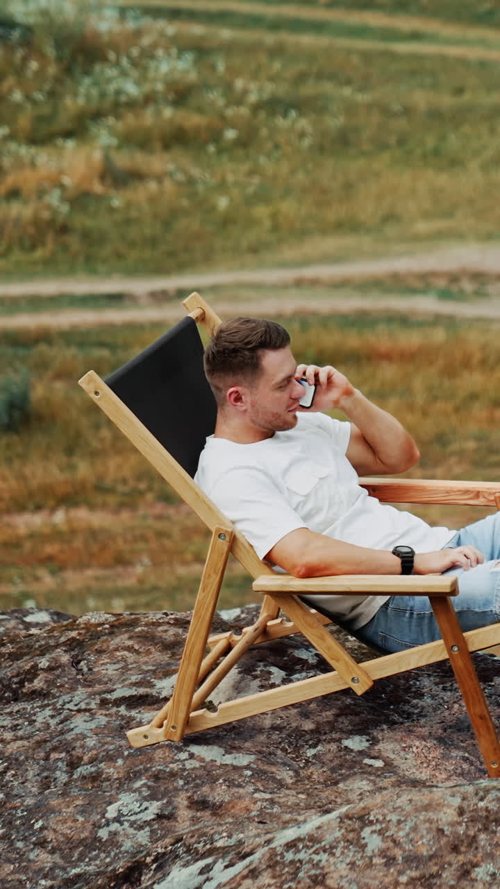 Man having rest in the nature. Caucasian male sits in a comfortable folding chair on the rock speaking on the phone. Top view. Vertical video