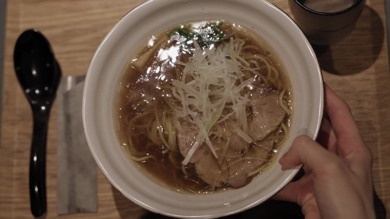 Mano che fa scivolare una ciotola di shoyu ramen con maiale chashu e porro su un tavolo di legno