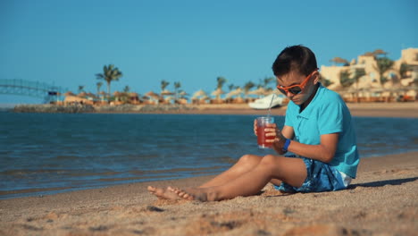 atractivo adolescente con gafas de sol bebiendo limonada en la playa. niño poniendo vidrio