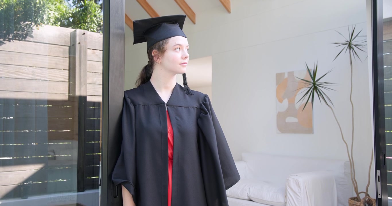 Donna adulta a casa in toga e tocco, che sposta lo sguardo e si dirige verso una porta aperta, preparandosi per la laurea