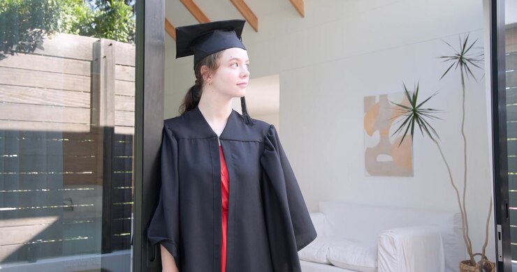 Donna adulta a casa in toga e tocco, che sposta lo sguardo e si dirige verso una porta aperta, preparandosi per la laurea