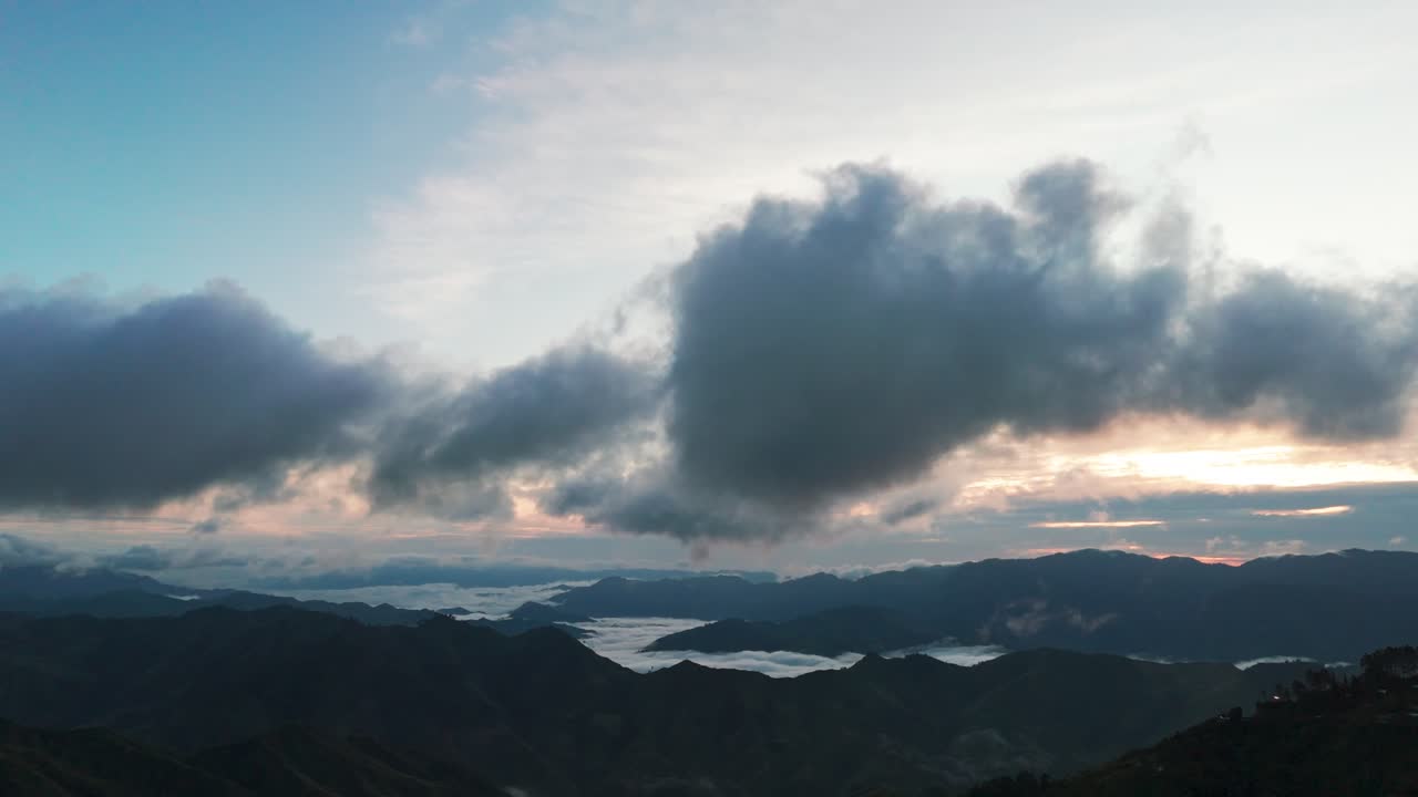 Wide mountain landscape at sunrise with dramatic low clouds and a sea of fog in the valleys, soft morning light and layered ridgelines under a blue sky, peaceful natural scenery