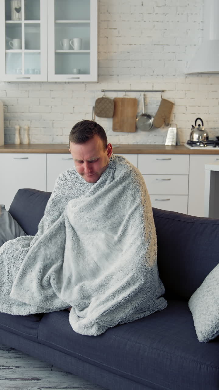 Man having fever. Young man feeling freezy and covering with warm blanket while sitting on a couch at home. Sick man wrapped with blanket during covid outbreak. Vertical video