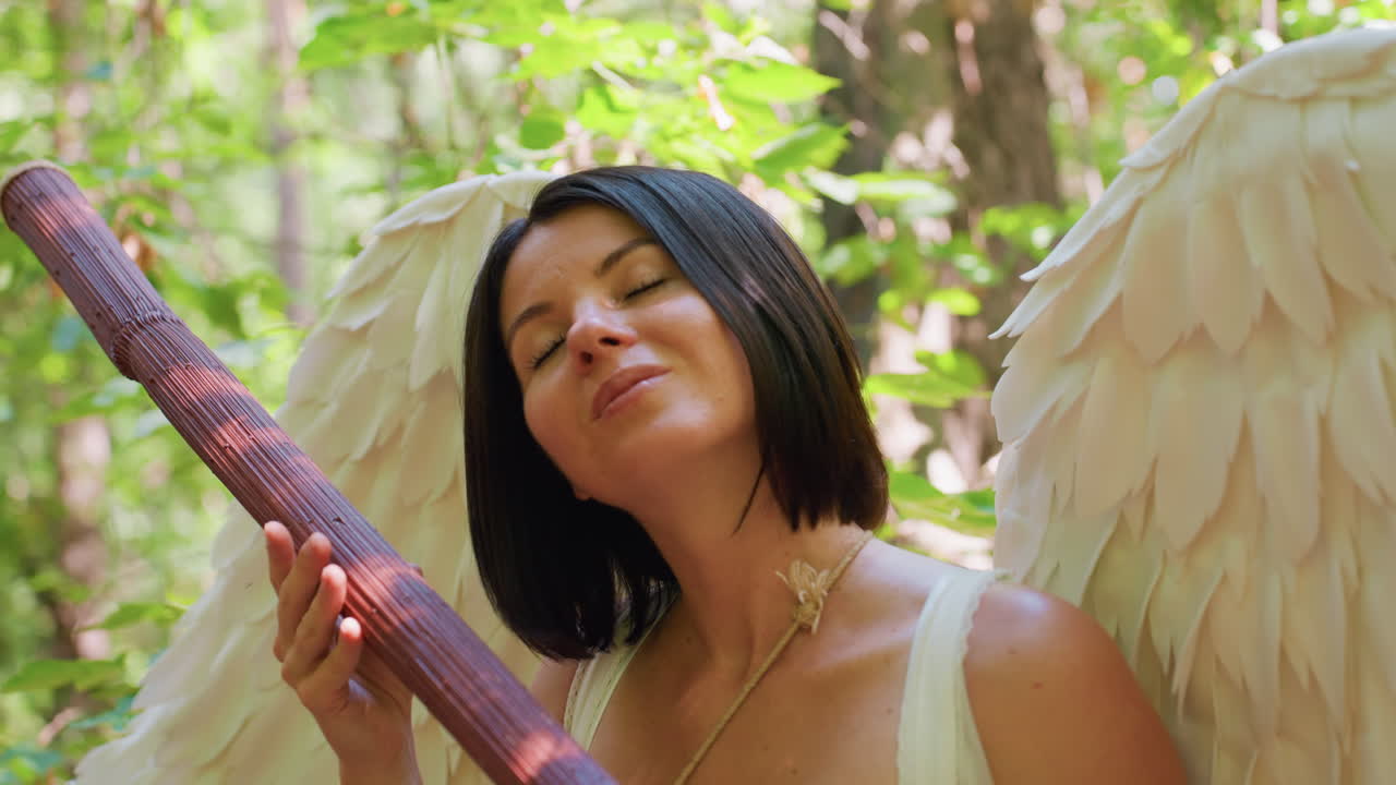 Serene angel woman with soft white wings holding wooden staff standing in lush green forest under warm sunlight expressing peace, inner strength, and divine calm in magical natural atmosphere