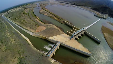 Top-Aerial-view-of-spillway-of-a-dam,-Beautiful-majestic-mountains-in-the-back-of-the-dam,-Sun-shines-in-the-green-water-of-the-dam