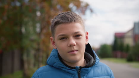 kid wearing blue jacket outdoors with a subtle smile and head tilted slightly, surrounded by vibrant autumn trees, blurred red-roofed house, and soft natural light