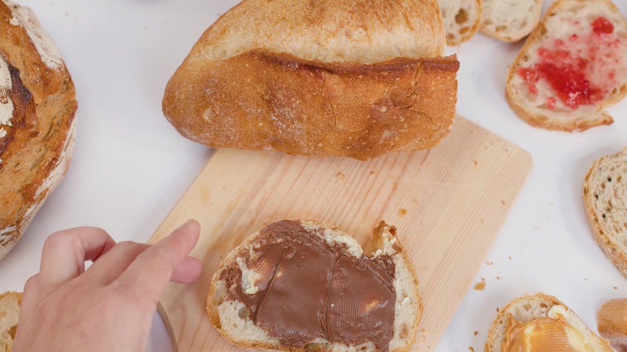 Hand spreads chocolate cream on crusty bread over a wooden board with white background.