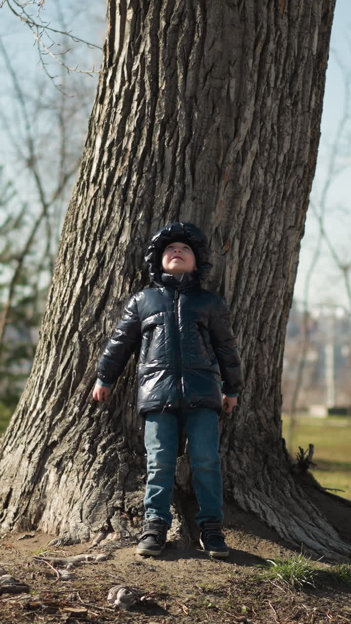 un niño joven se apoya de espaldas contra un gran tronco de árbol, mirando hacia arriba con los brazos extendidos contra el árbol, está vestido cálidamente con una chaqueta negra brillante, vaqueros y botas, con árboles circundantes