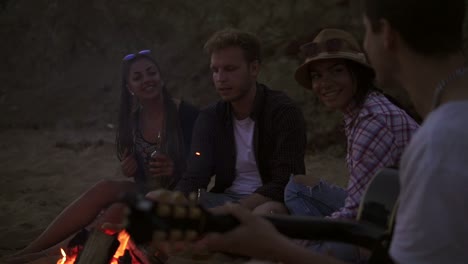 Picnic-of-young-people-with-bonfire-on-the-beach-in-the-evening.-Cheerful-friends-singing-songs-and-playing-guitar.-Slow-Motion