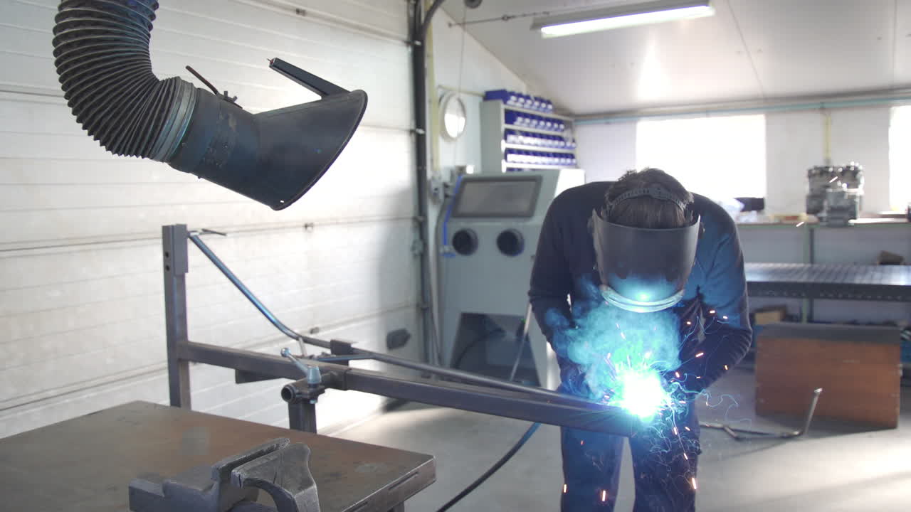 산업 작업 공간에서 작업하는 용접공은 녹색 불꽃과 스파크가 있는 금속 구조물을 만들고 오버헤드는 통풍과 배기관을 보여줍니다.