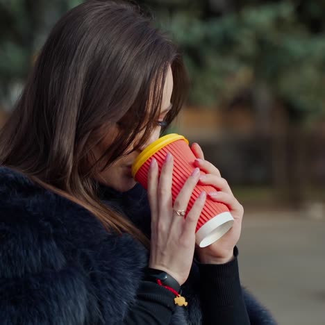 long-haired girl with plastic cup outdoors. attractive young woman in fur coat enjoys drinking coffee in the city park. slow motion.