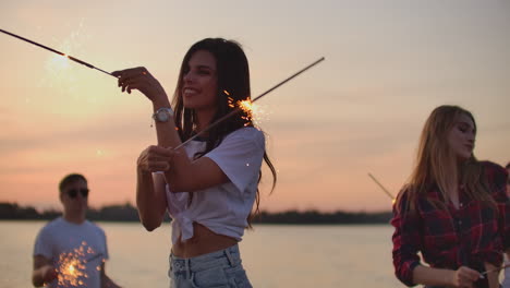 The-group-of-young-friends-is-dancing-with-big-bengal-lights-on-the-sand-coast.-This-is-cool-theamy-summer-evening-on-the-open-air-party.