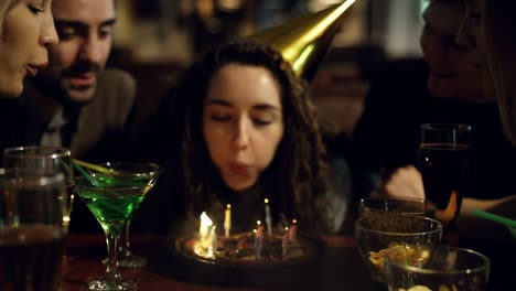 close-up shot of young pretty woman blowing out candles on birthday cake. happy people are laughing and clapping hands.