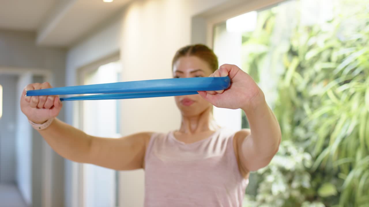 Video verticale: Donna in top rosa che tiene una fascia blu e si allena in sala, con orologio visibile.