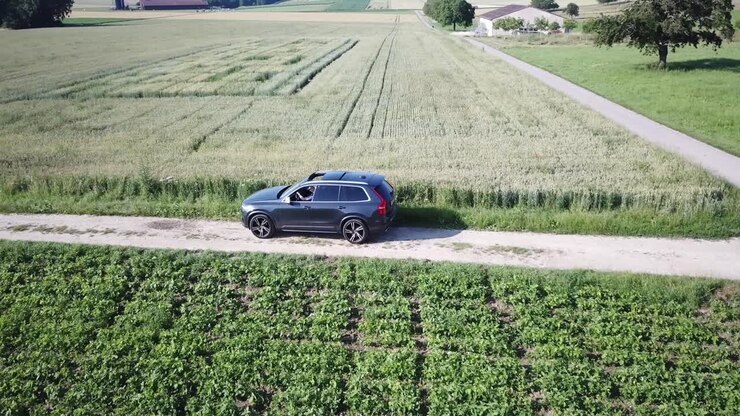 Un'auto Volvo XC90 SUV procede lentamente su una piccola strada dissestata tra campi agricoli, dopo un incrocio tra due strade, nella campagna svizzera, Vaud; ripresa aerea con drone.