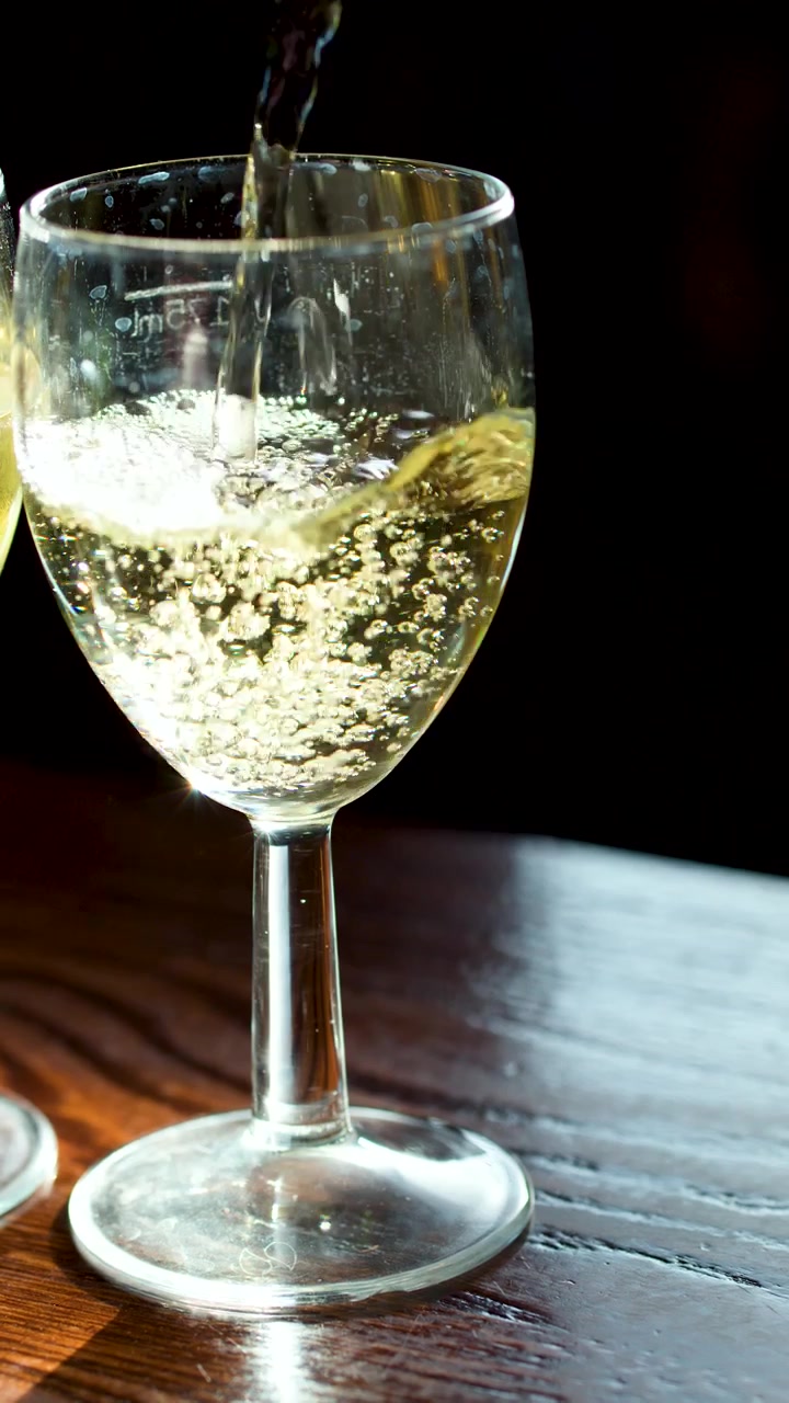 Vino bianco frizzante versato in un bicchiere trasparente, bollicine che salgono, luce solare naturale, tavolo di legno rustico