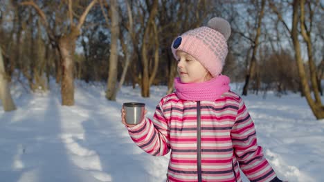 Smiling-child-kid-girl-drinking-hot-drink-tea-from-cup,-trying-to-keep-warm-in-winter-park-forest