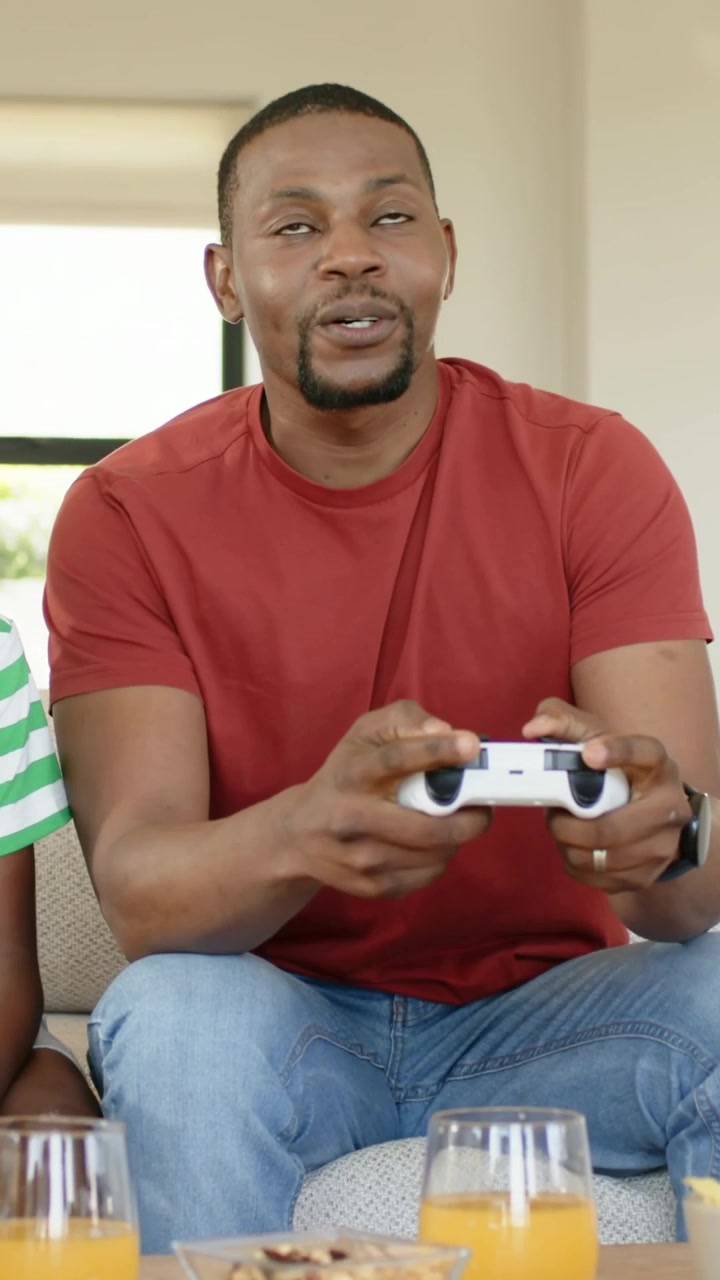 Vertical video: Turning to offscreen cue, dad in red tee grabbing controllers at home, son joining